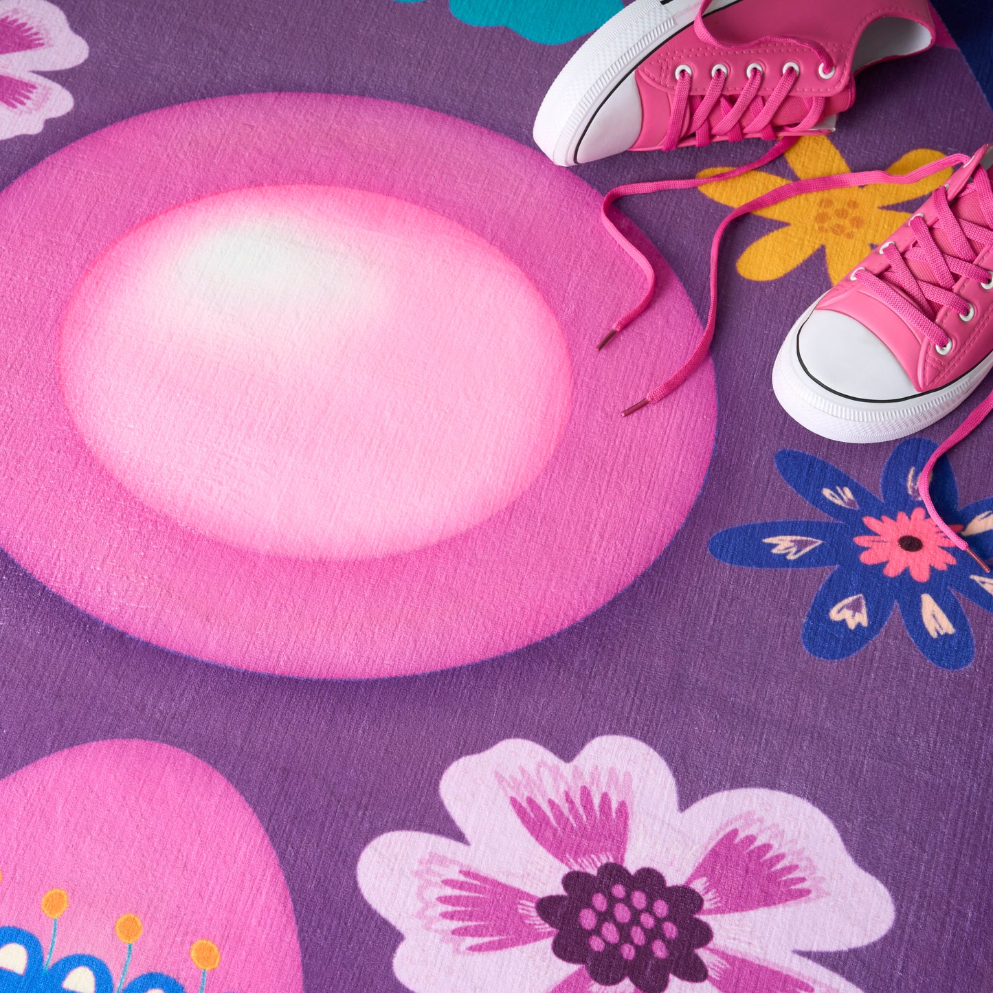 Pink round plush play mat on a purple floral rug, with pink canvas sneakers and white toe caps nearby.
