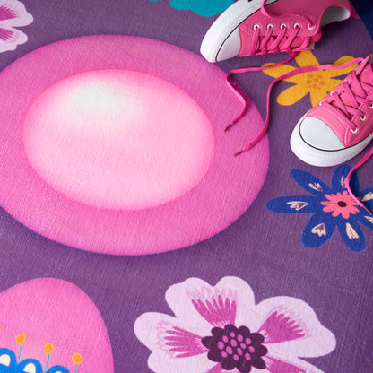 Pink round plush play mat on a purple floral rug, with pink canvas sneakers and white toe caps nearby.