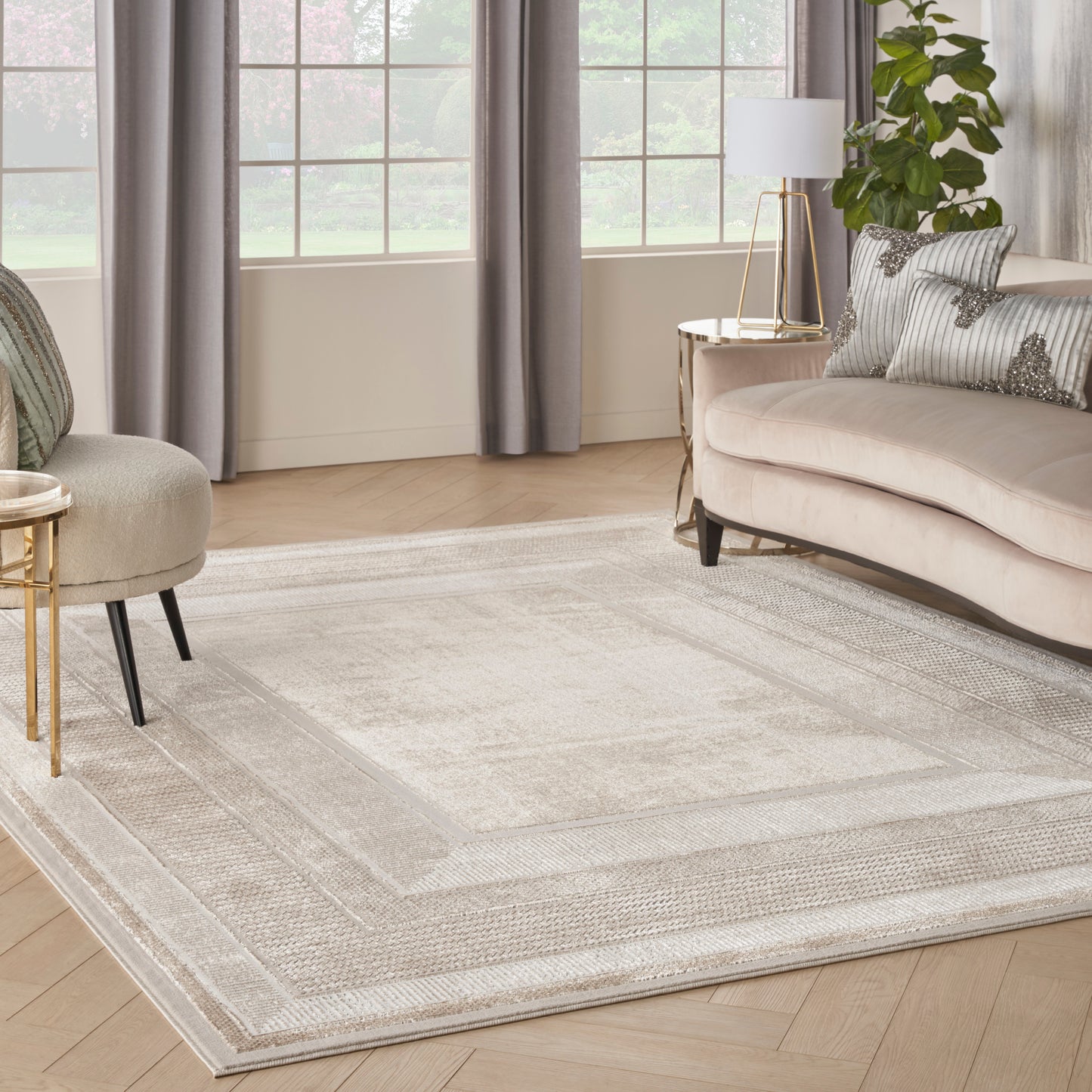 Light gray rectangular area rug with a bordered design in a bright living room; beige curved sofa, gold side table, plant.