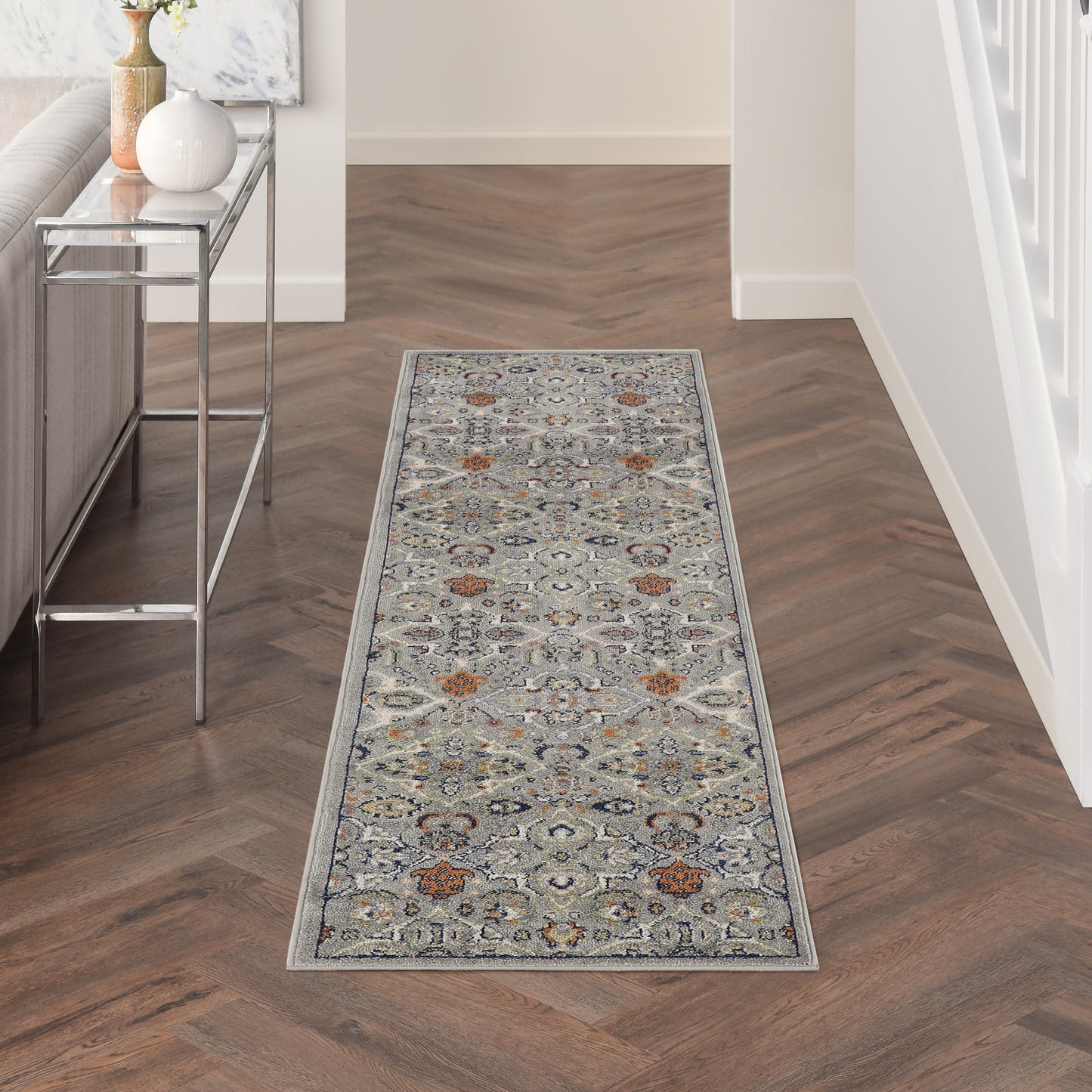 Long narrow Persian-inspired gray-blue hallway runner with orange motifs on a dark wood herringbone floor.