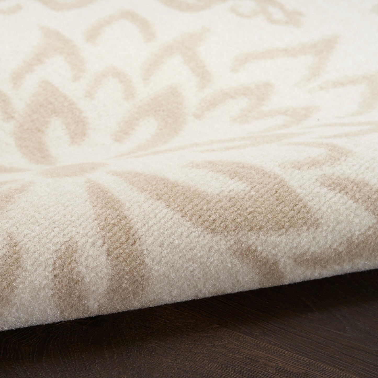 Cream-beige patterned area rug with soft plush pile, featuring leaf-like motifs on a dark wood floor.
