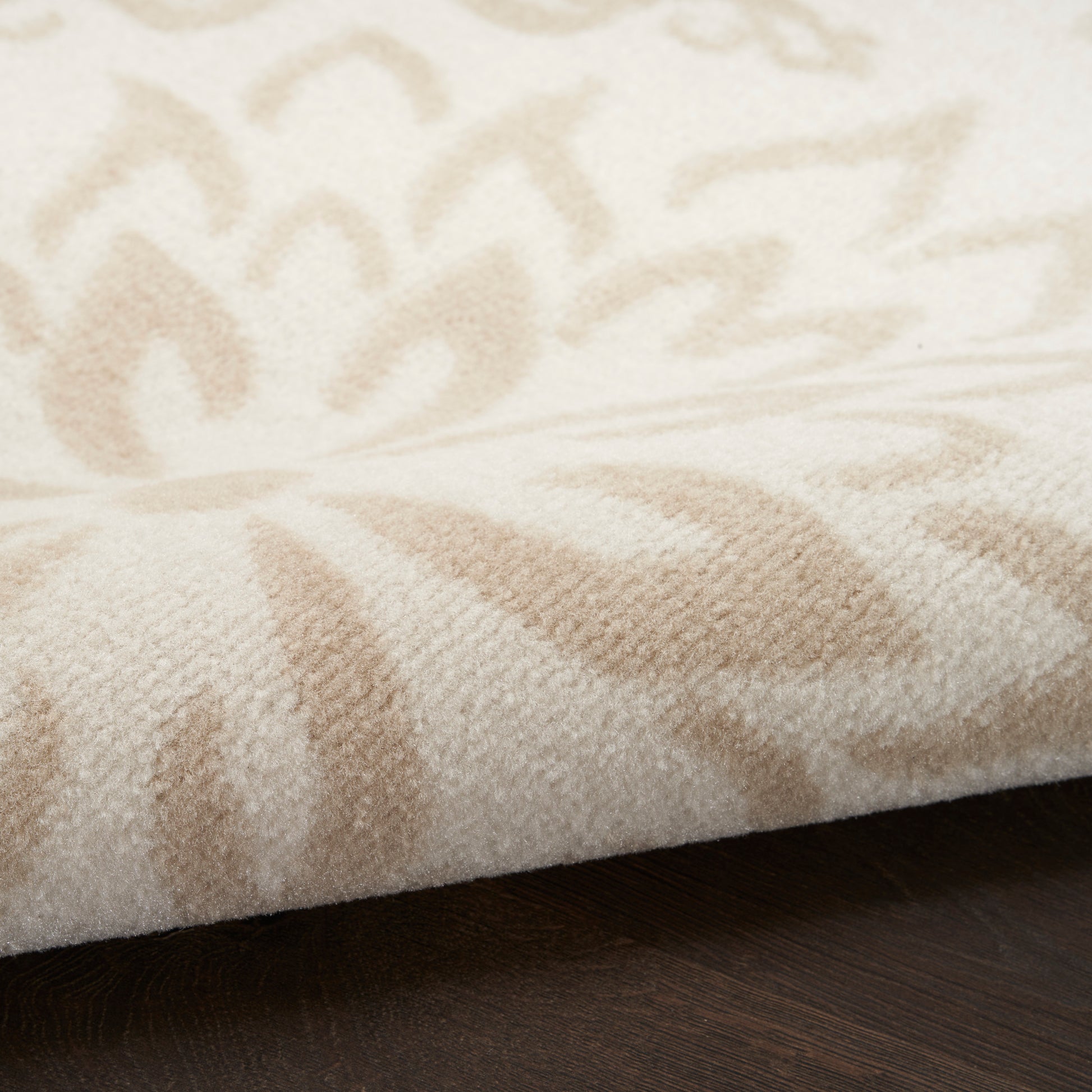Cream-beige patterned area rug with soft plush pile, featuring leaf-like motifs on a dark wood floor.