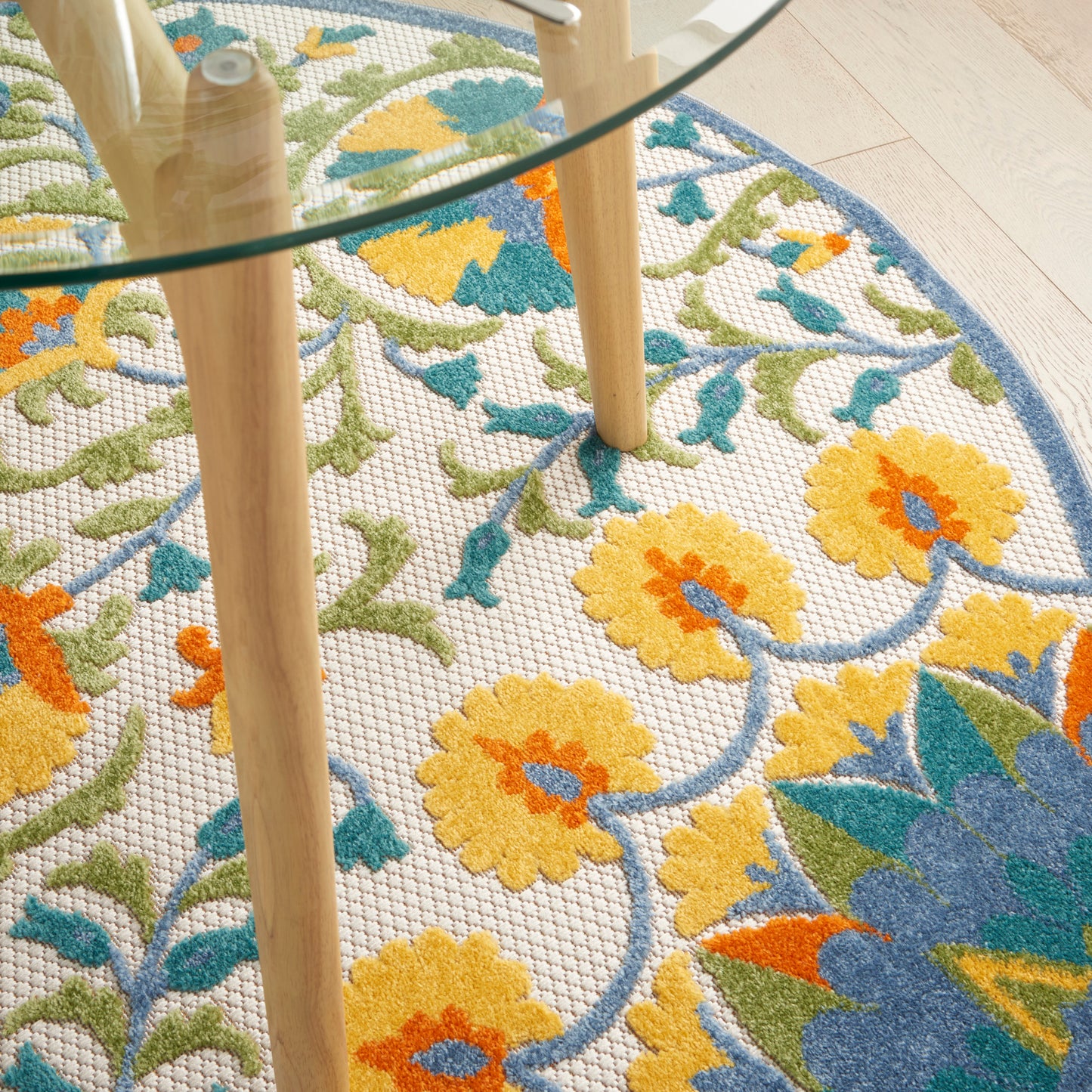 Round glass coffee table with light wood legs on a colorful floral rug featuring yellow flowers and blue-green leaves.