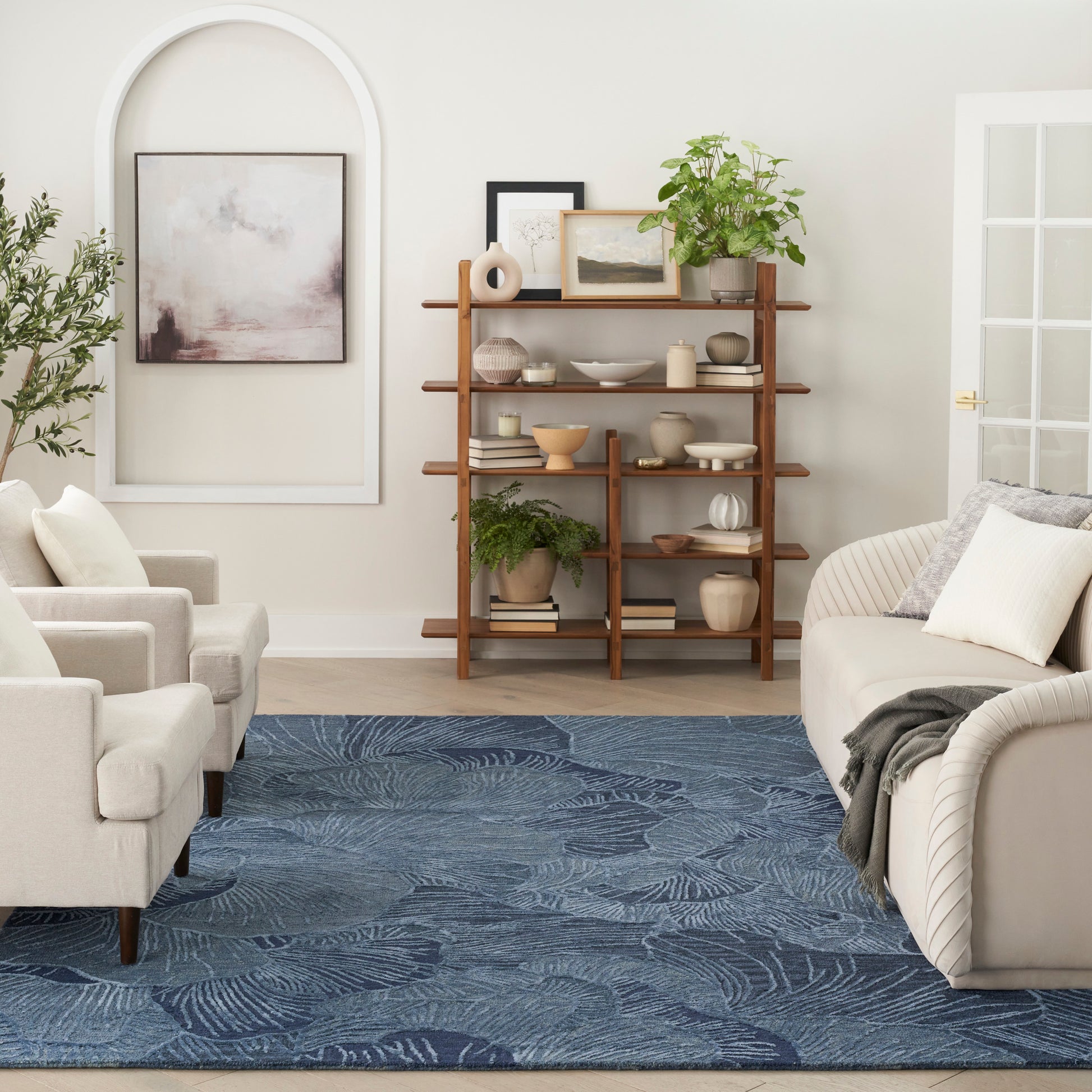 Living room with a wooden open bookshelf, beige sofas, blue palm-pattern rug, potted plants, arched decor & framed art.
