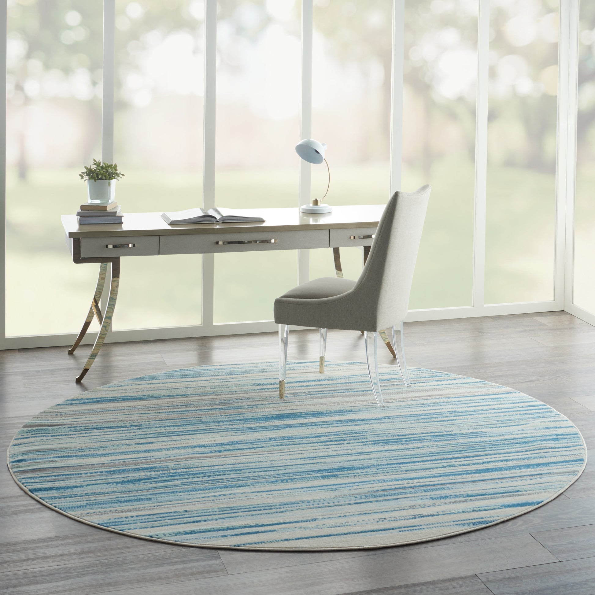 Round blue striped rug under a white modern desk with gold curved legs and a grey upholstered chair in a sunlit home office.