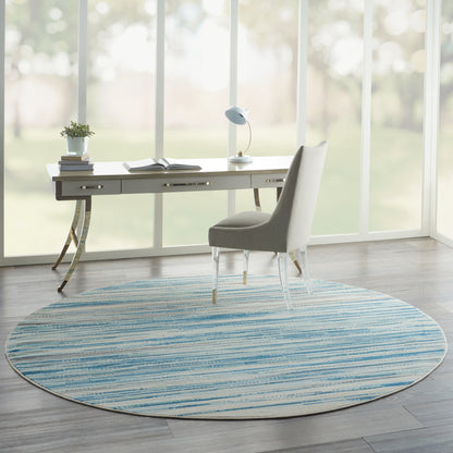 Round blue striped rug under a white modern desk with gold curved legs and a grey upholstered chair in a sunlit home office.