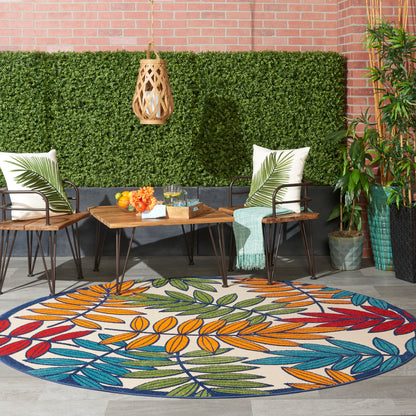 Outdoor patio with round leaf rug, wicker chairs, wooden table, tropical cushions, hanging lantern, plants, brick wall.
