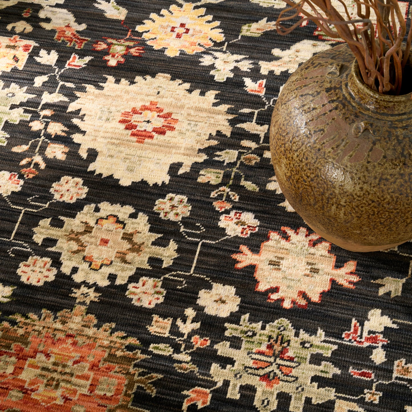 Dark patterned rug with ivory floral diamond motifs and red accents, topped by a brown ceramic vase with dried branches.