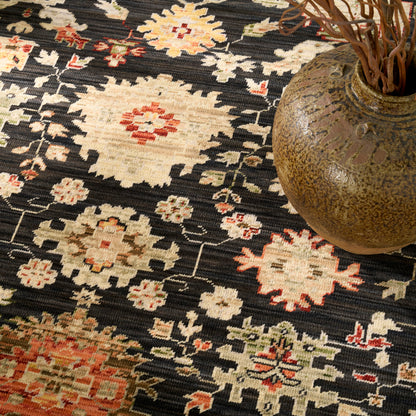 Dark patterned rug with ivory floral diamond motifs and red accents, topped by a brown ceramic vase with dried branches.