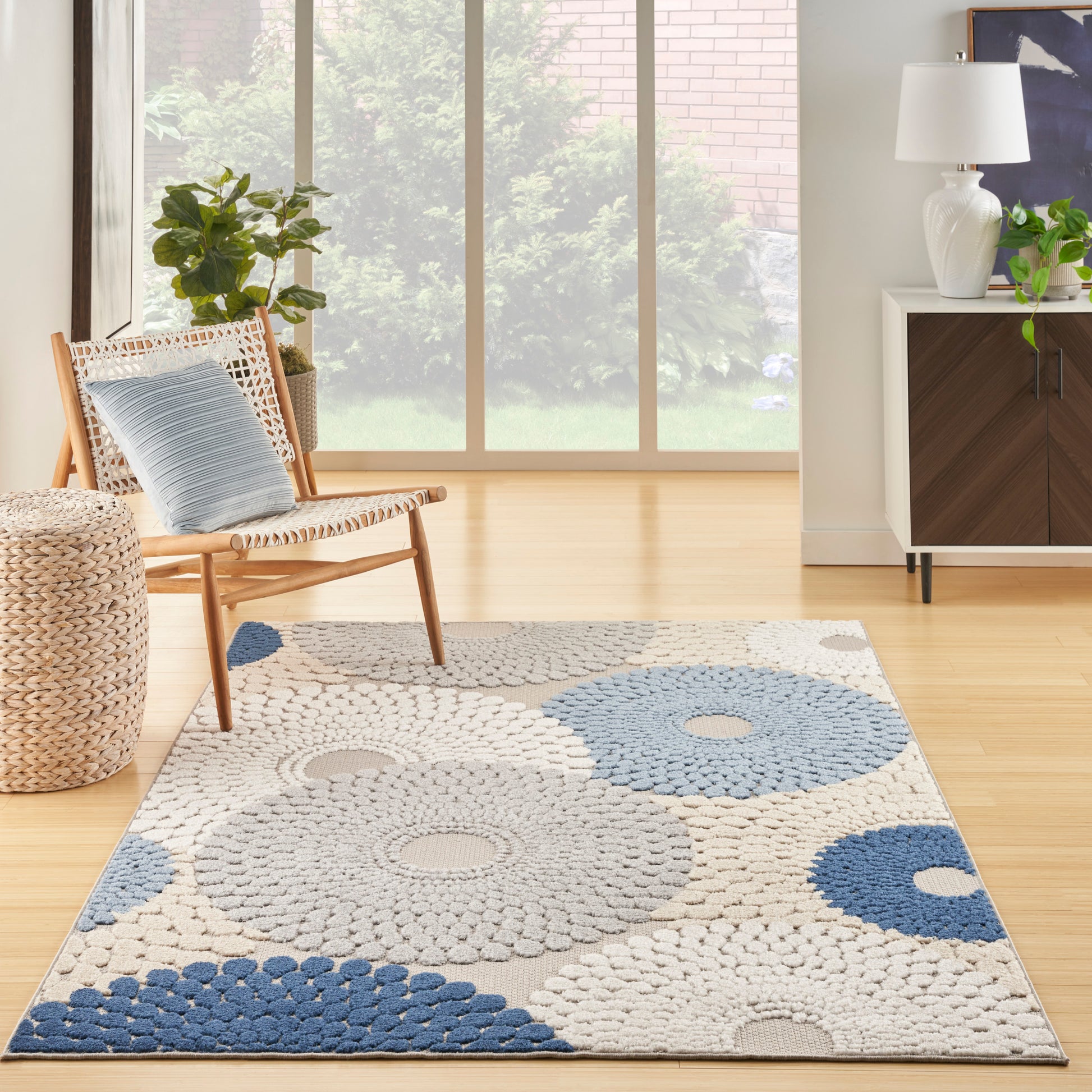 Bright living room with large windows, woven wood chair, rectangular blue and beige rug, wicker basket, plants, and a modern sideboard.