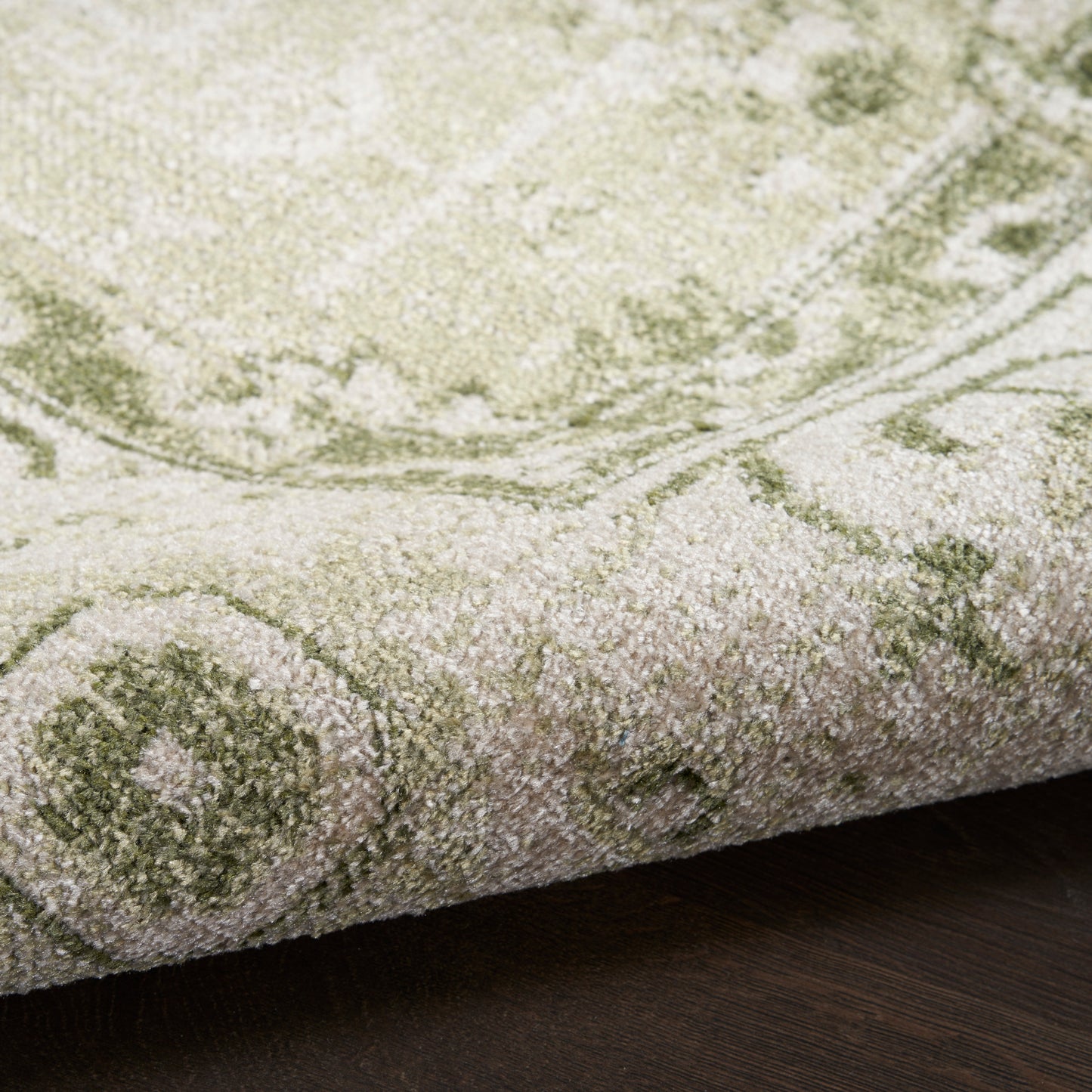 Cream and sage-green patterned rug with plush pile, rolled edge on dark wood floor.