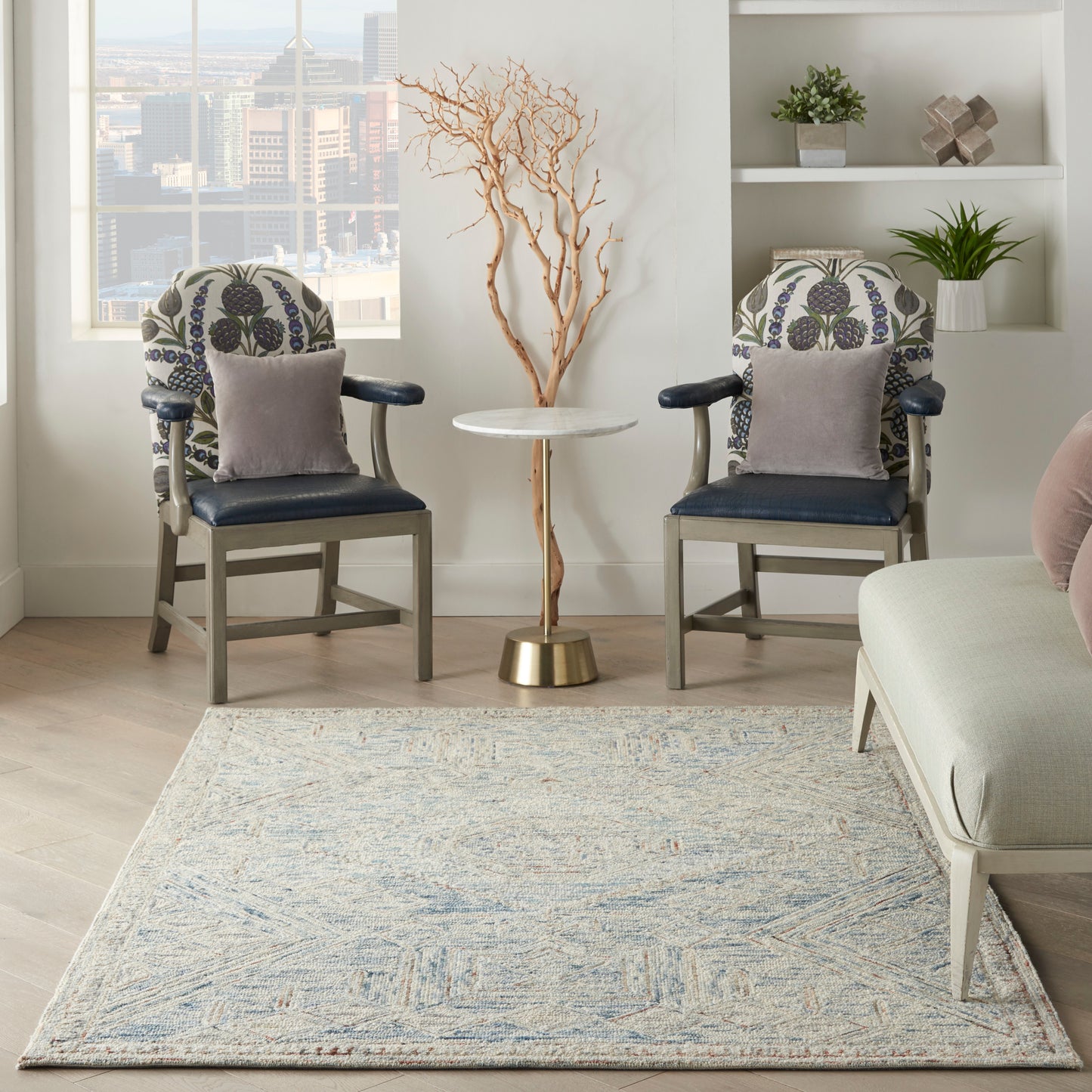 Two floral upholstered chairs with navy seats and gray cushions flank a marble-top table; dried tree sculpture and blue rug.