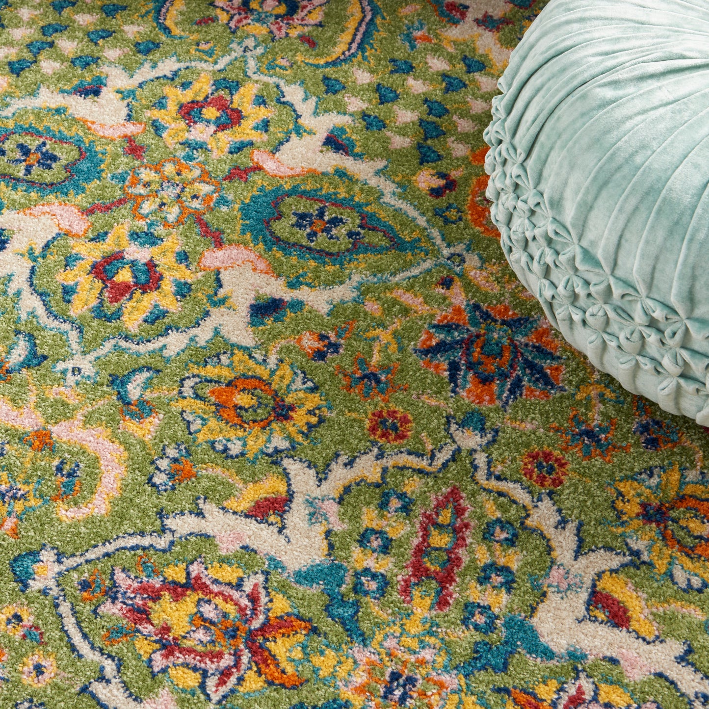 Colorful Persian-style rug with green, blue, orange and pink floral motifs; teal round pleated velvet pouf resting on it.