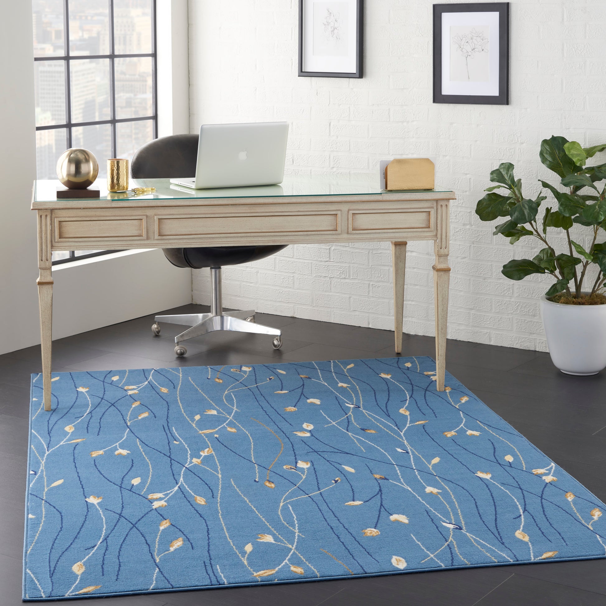 Light wood desk with glass top, laptop, black rolling chair, blue patterned rug, white brick wall with frames, plant.