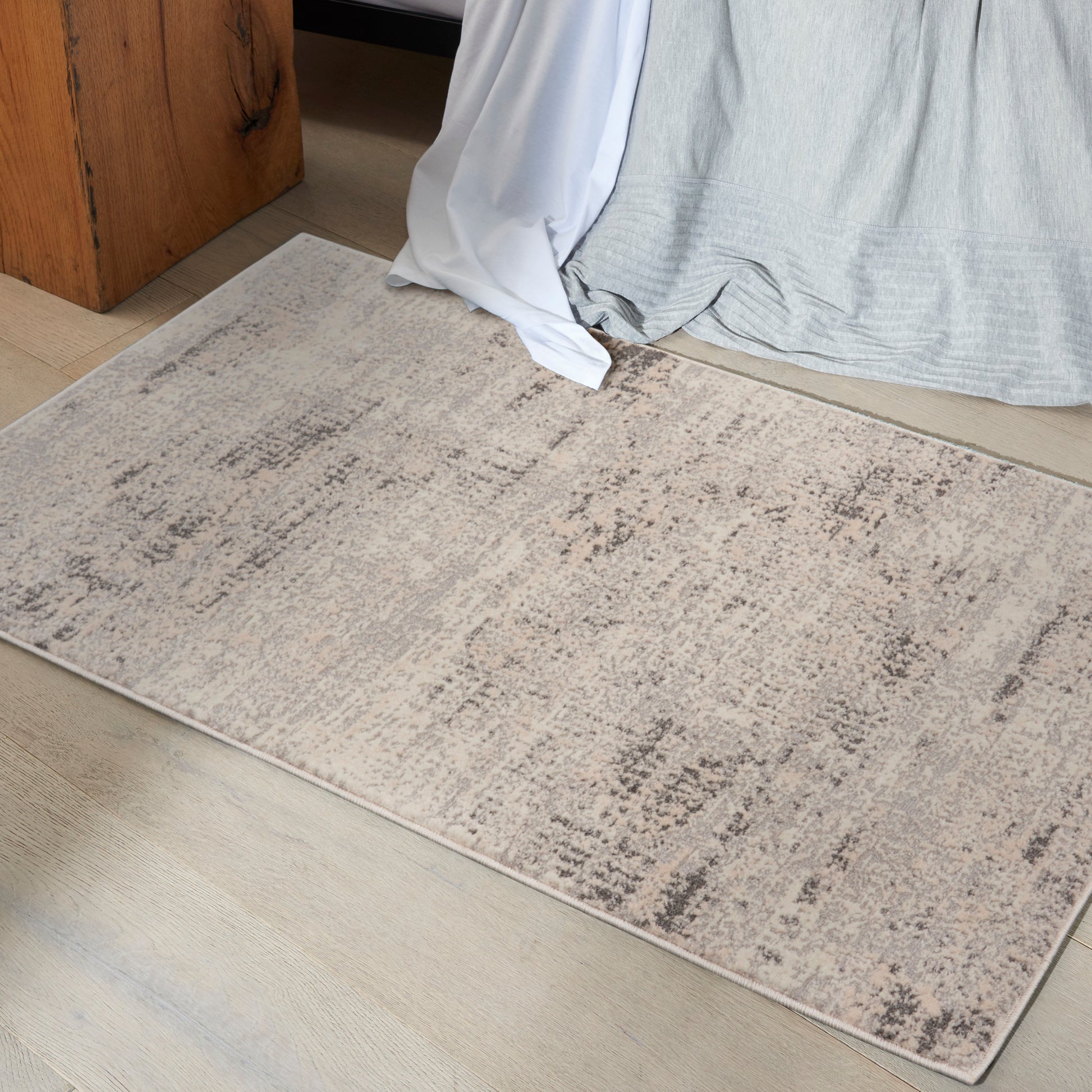Neutral beige rectangular area rug with a subtle distressed taupe pattern on a light wood floor beside a bed.