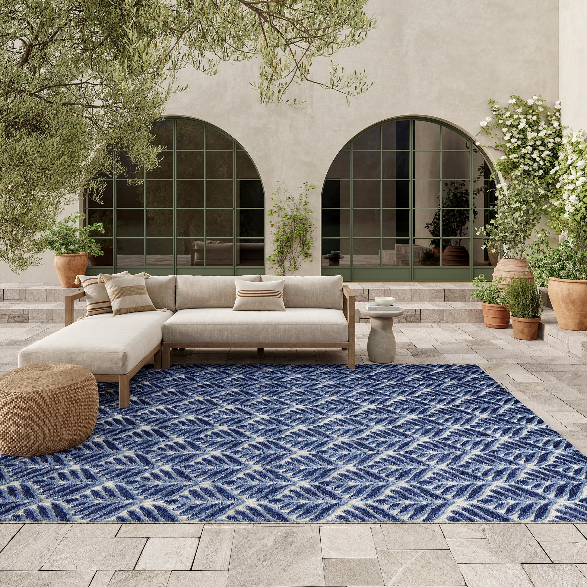 Outdoor patio with beige sectional sofa on a blue patterned rug, olive trees, potted plants, and arched green-framed windows.