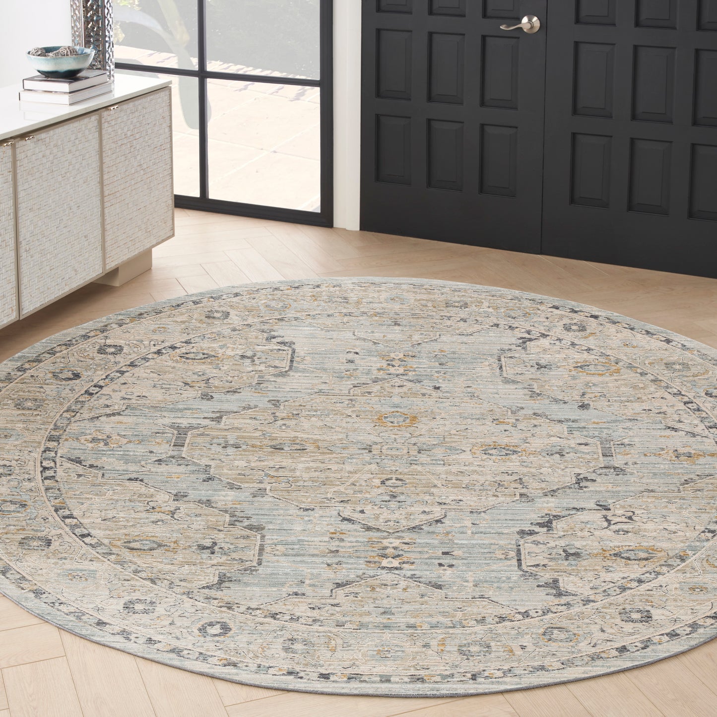 Round traditional rug in muted blue, beige and ivory on light wood flooring beside a black paneled door.