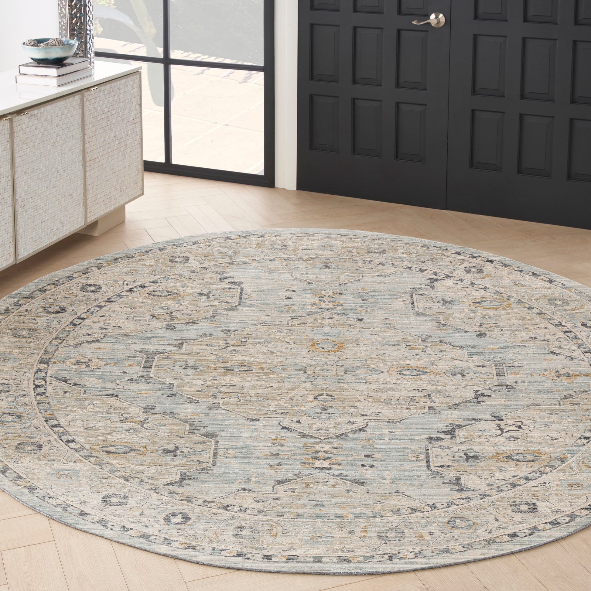 Round traditional rug in muted blue, beige and ivory on light wood flooring beside a black paneled door.