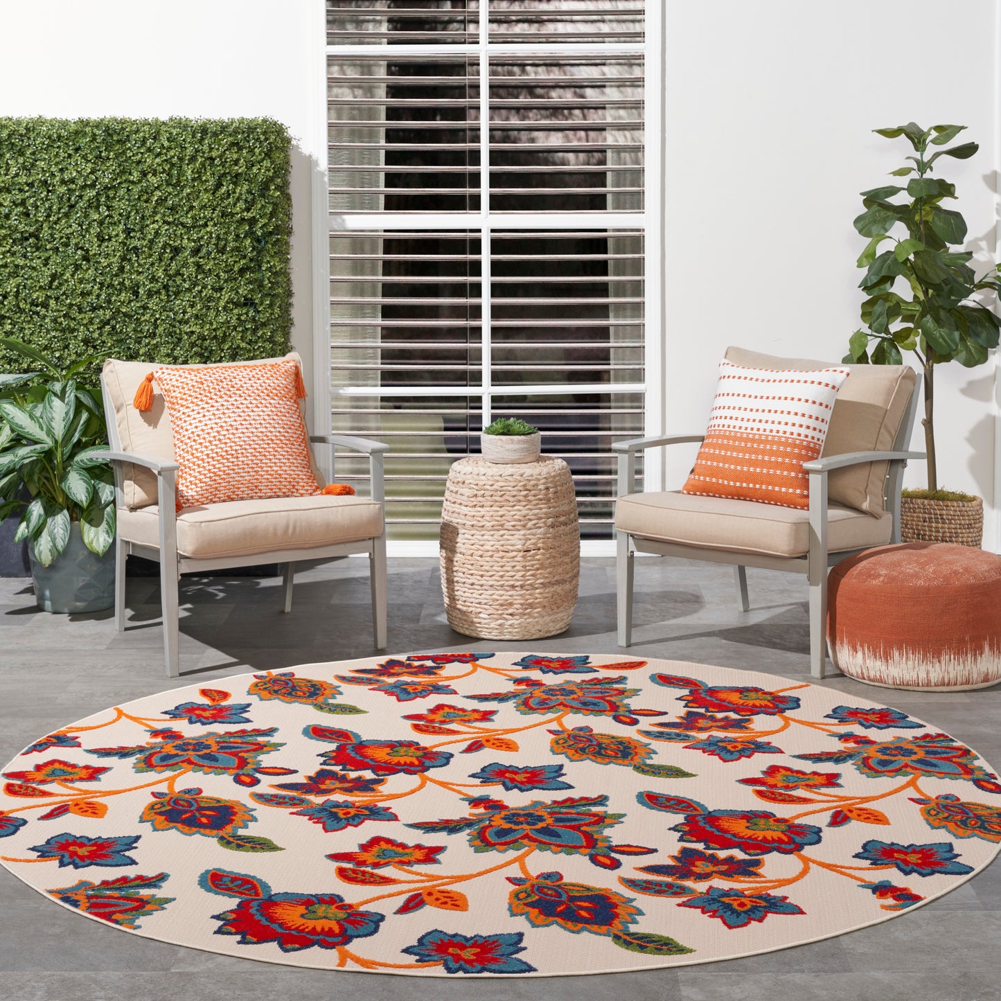 Round rug with bright orange, red & blue florals; two patio chairs with orange cushions, woven side table.