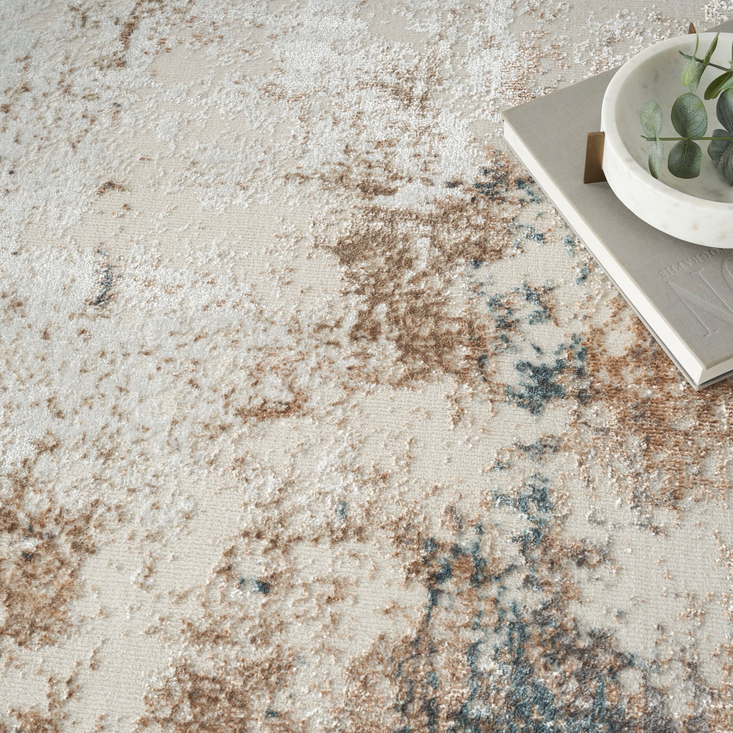 Abstract beige rug with brown and blue marbling; corner features a white planter on a gray tray with a book.