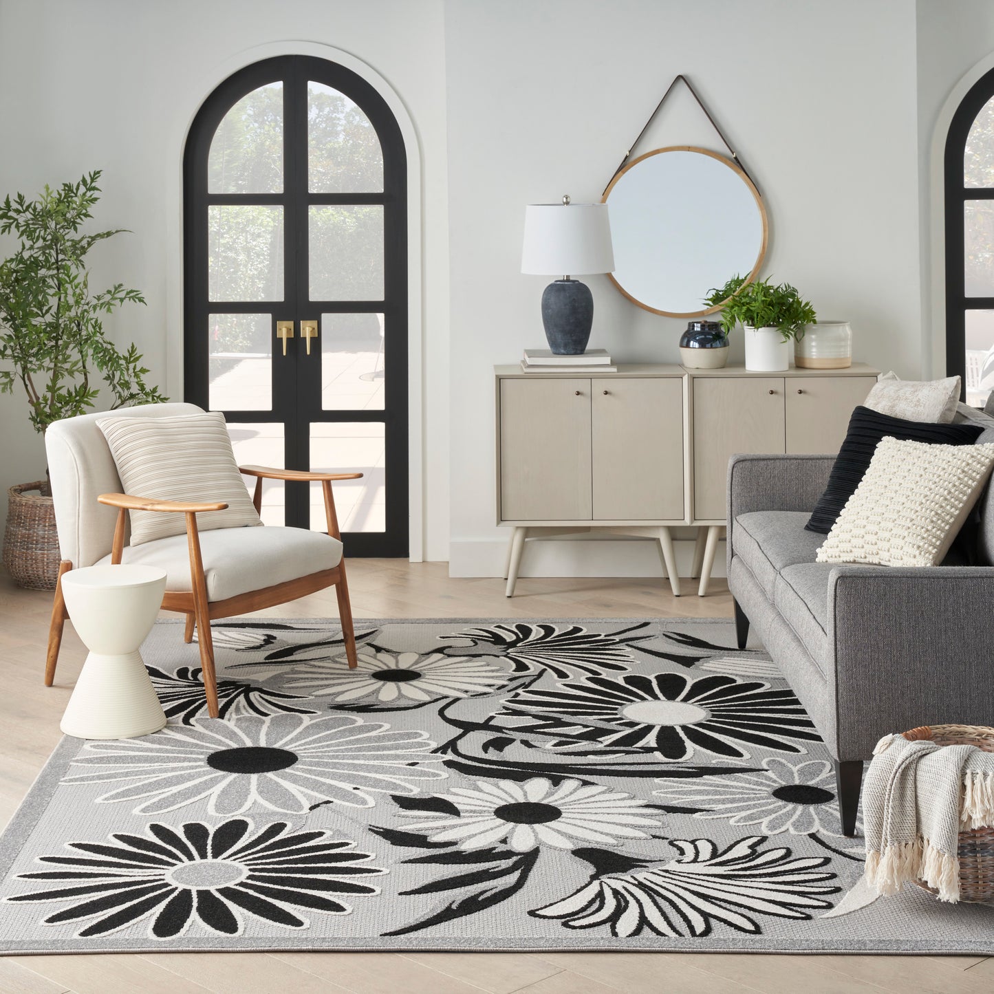 Living room with a large black-and-white floral rug, gray sofa, wood armchair, round mirror, and arched black doors.