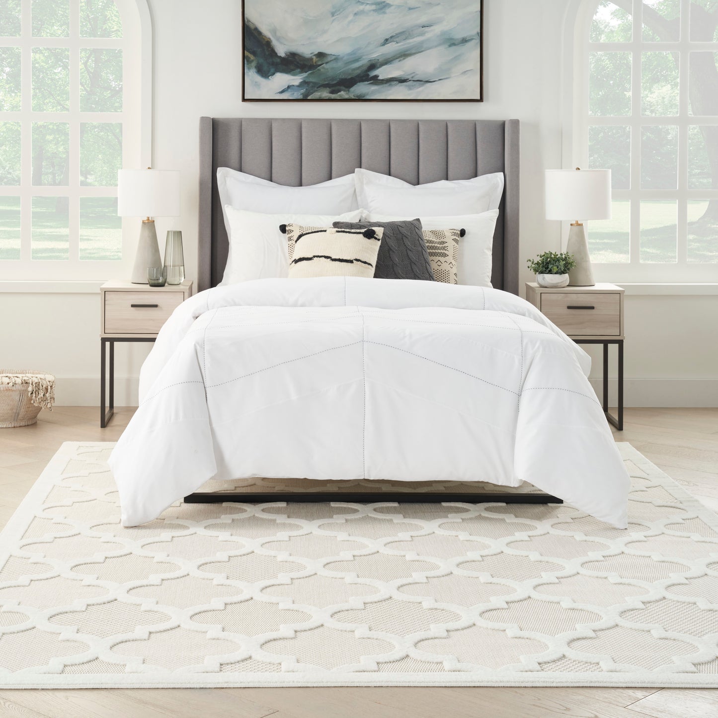 White bedding on a gray tufted headboard bed with decorative pillows, beige nightstands, lamps, and a patterned cream rug.