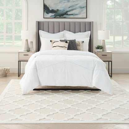 White bedding on a gray tufted headboard bed with decorative pillows, beige nightstands, lamps, and a patterned cream rug.