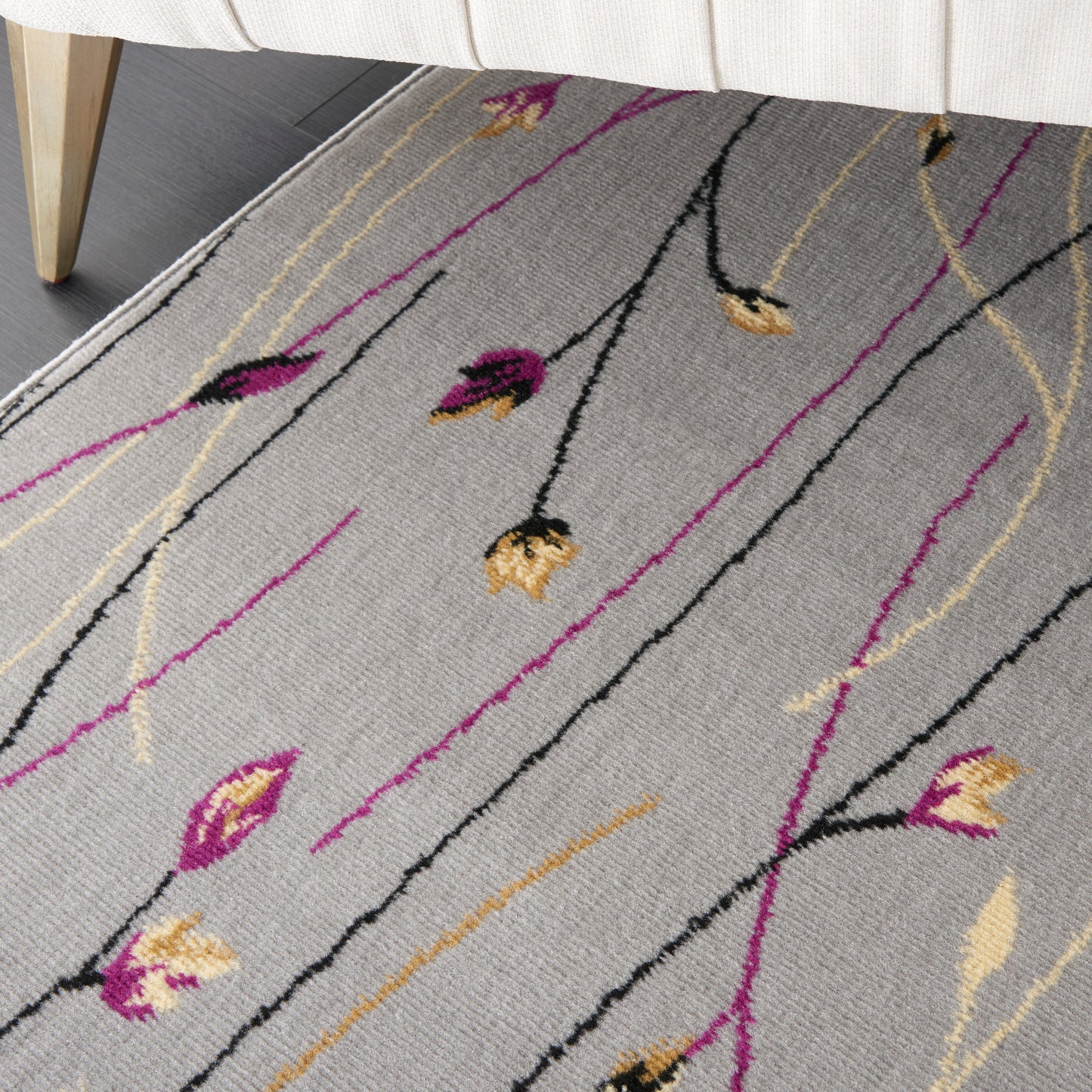 Light gray rug with slender black, magenta and gold floral lines and buds, under a beige sofa.
