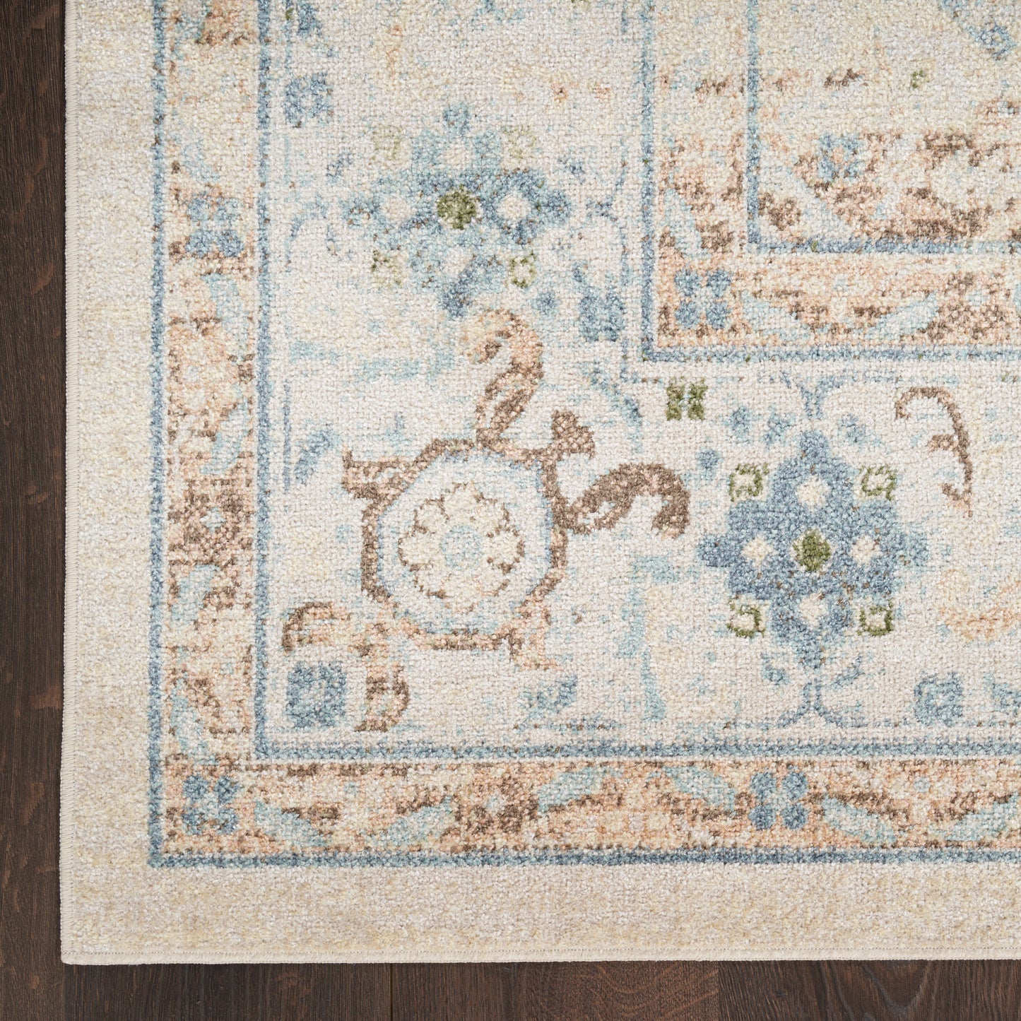 Corner view of a pale beige rug with blue floral border and medallion on dark wood floor.