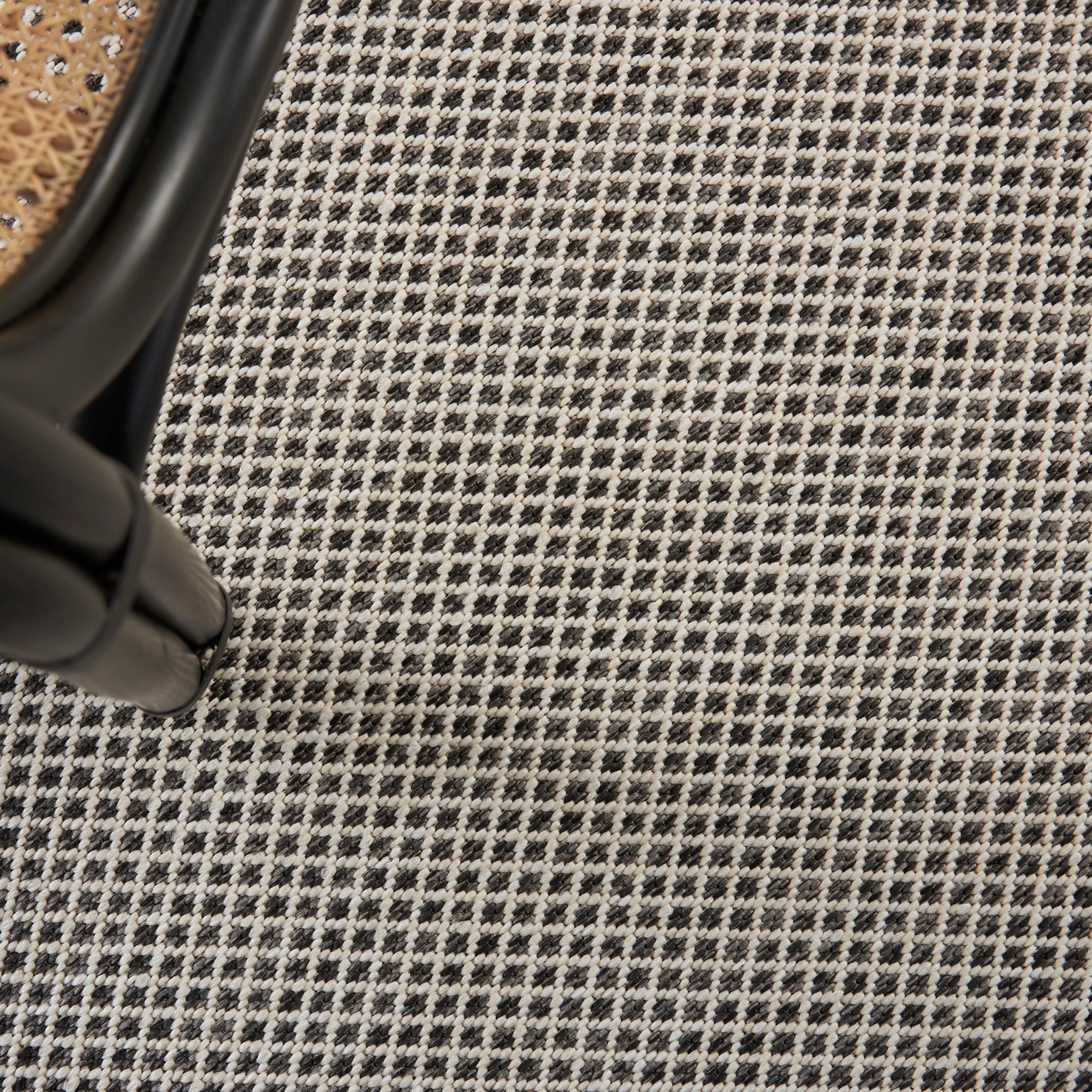 Black metal chair leg with curved joint beside a woven rattan seat on a black-and-white checkered textile rug