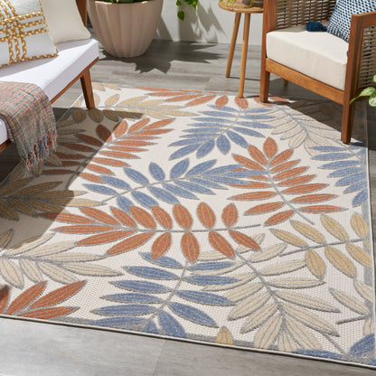 Outdoor woven rug with orange, blue and beige leaf pattern; pairs with white-cushioned seating and wooden furniture.