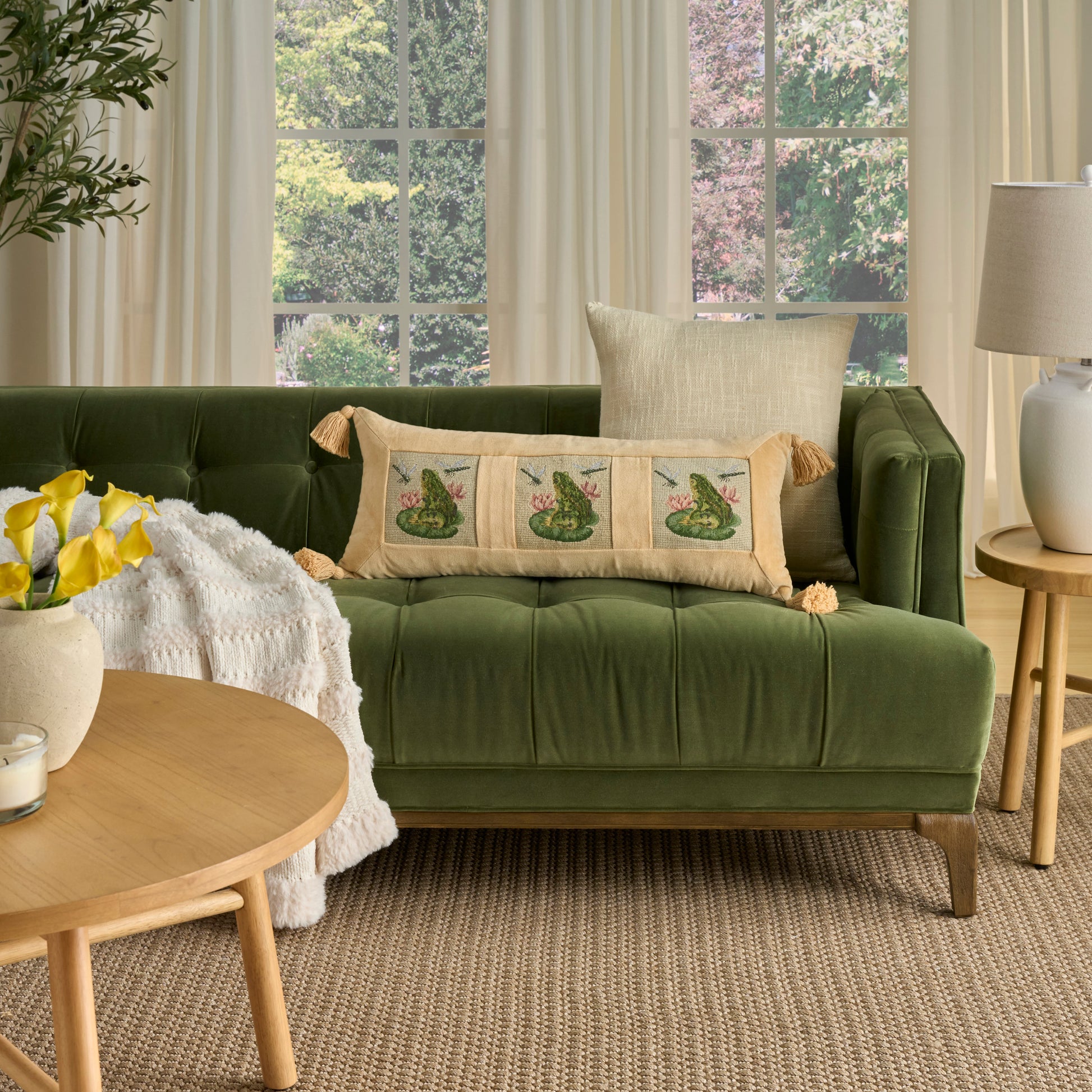 Olive green velvet tufted sofa with beige botanical cushions & cream throw, round wood coffee table, sunlit living room.
