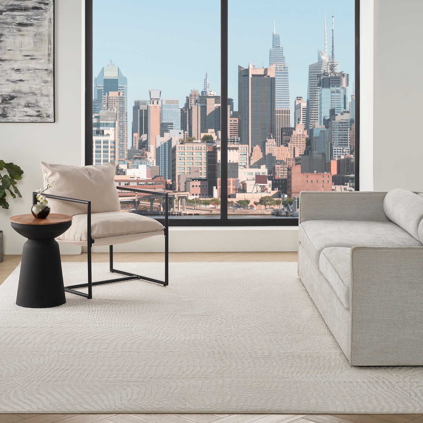 Bright modern living room with a city skyline view, beige sofa, metal-framed armchair, and round black side table on a rug.