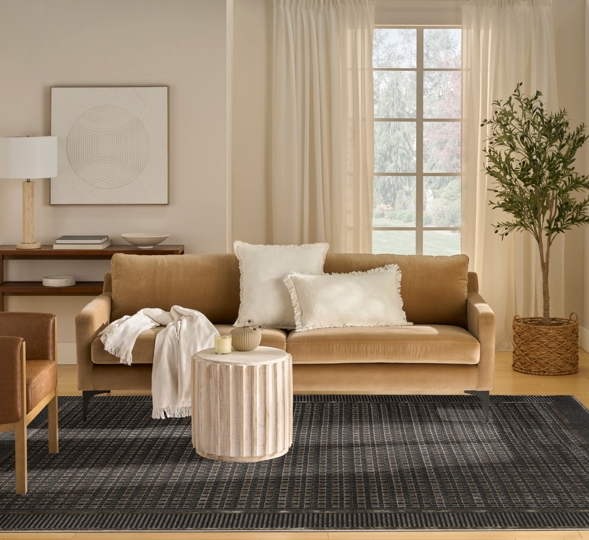 Tan sofa with white pillows, cream throw; round ribbed wood drum table, dark geometric rug, plant in wicker basket.