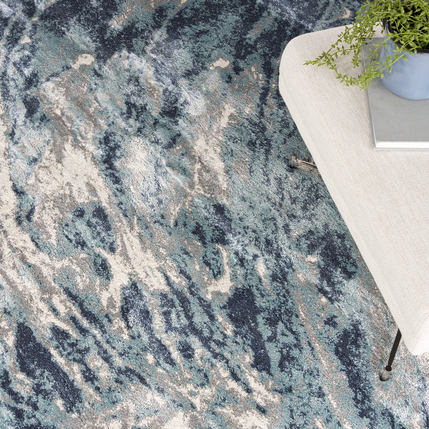 Abstract blue-gray area rug with ivory streaks; modern low-pile style beside a light wood table with a potted plant.