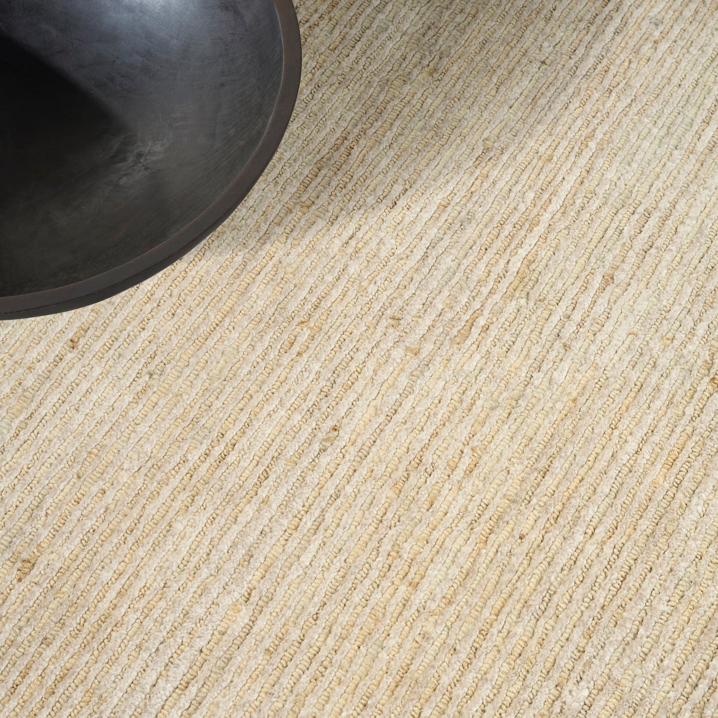 Beige natural-fiber rug with striped weave; dark round bowl or planter partially visible in top-left.