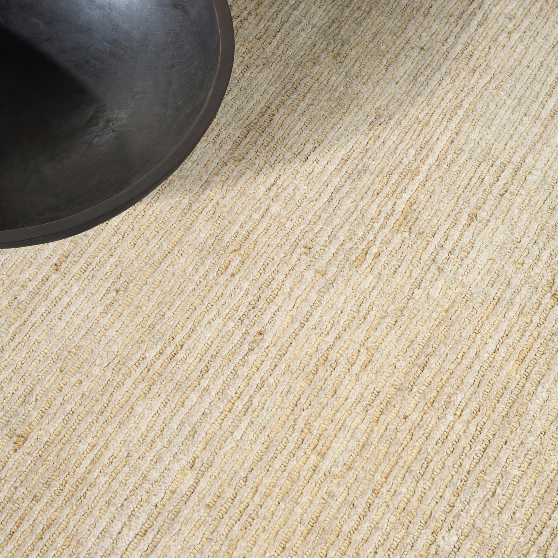 Beige natural-fiber rug with striped weave; dark round bowl or planter partially visible in top-left.