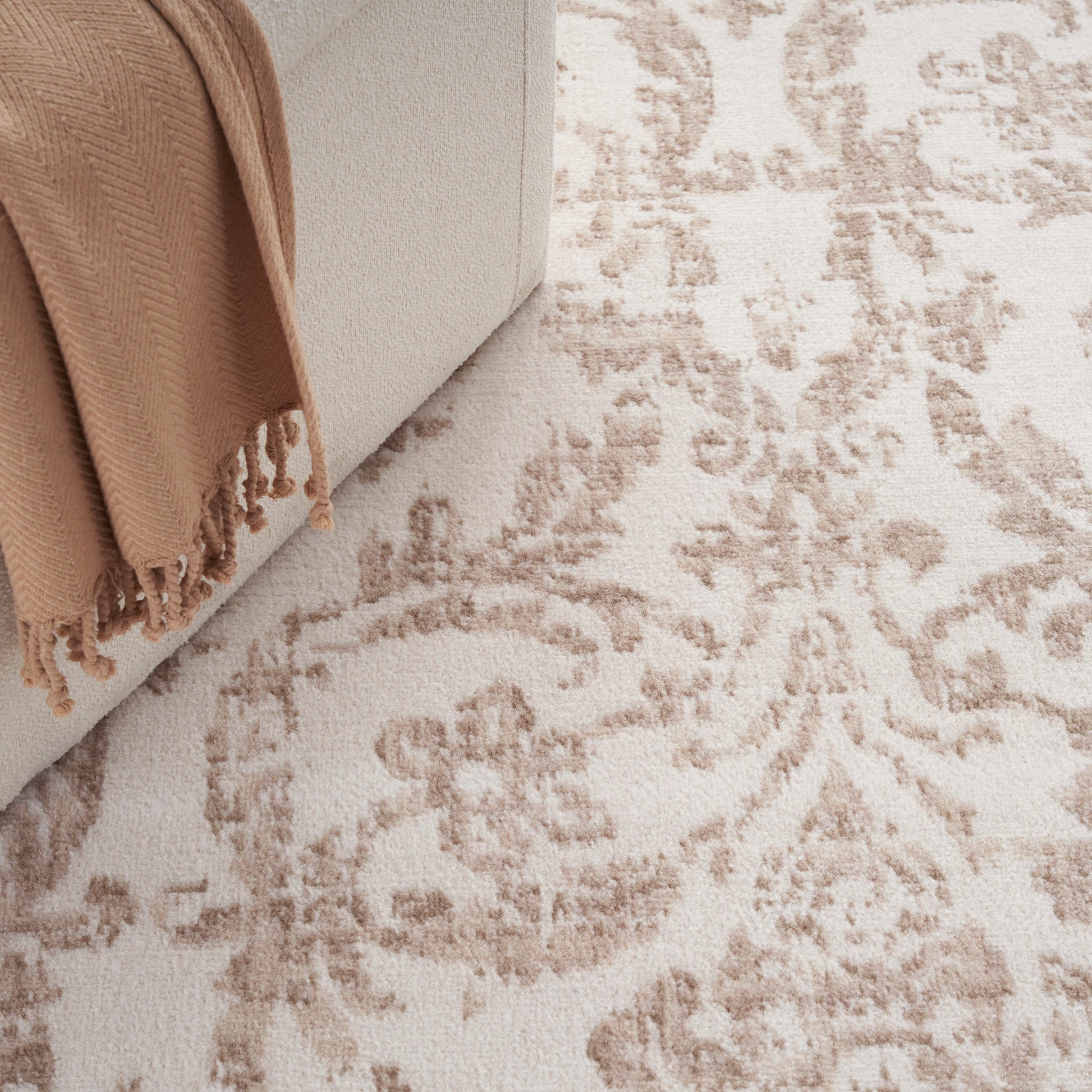 Beige sofa with tan fringed throw draped over the arm, above a cream patterned rug.