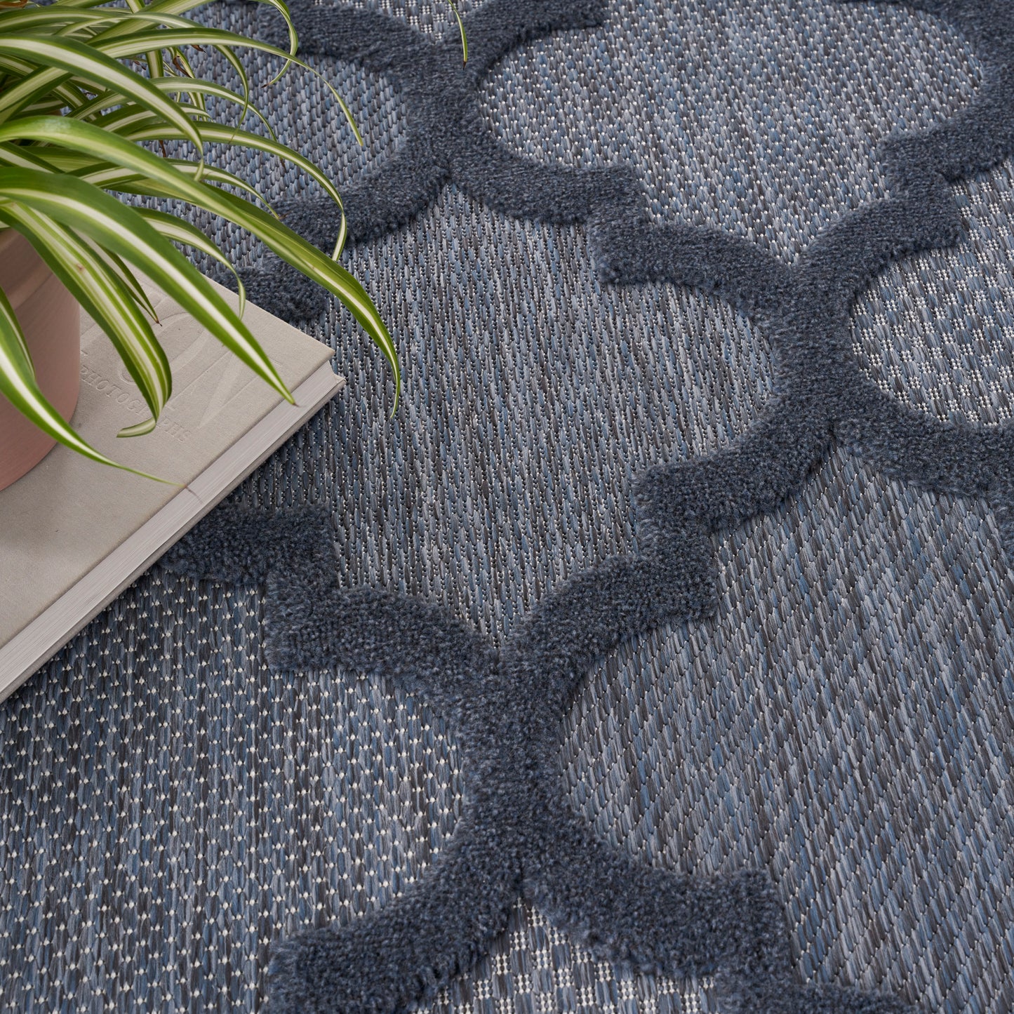 Blue woven rug with dark navy interlocking quatrefoil motifs and textured base; potted plant and magazines nearby.