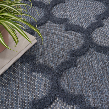 Blue woven rug with dark navy interlocking quatrefoil motifs and textured base; potted plant and magazines nearby.