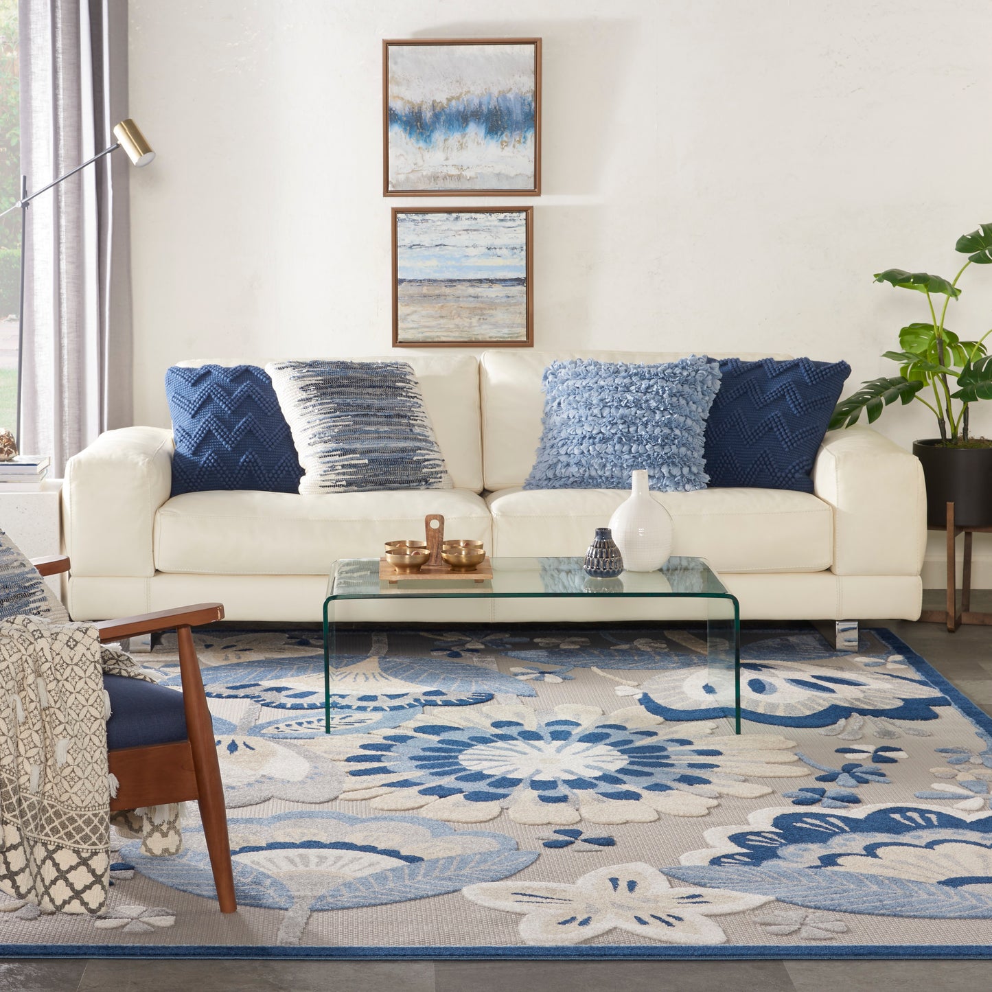 Cream sofa with blue cushions, glass coffee table, blue floral rug, wooden chair, floor lamp, and a plant in a coastal home.