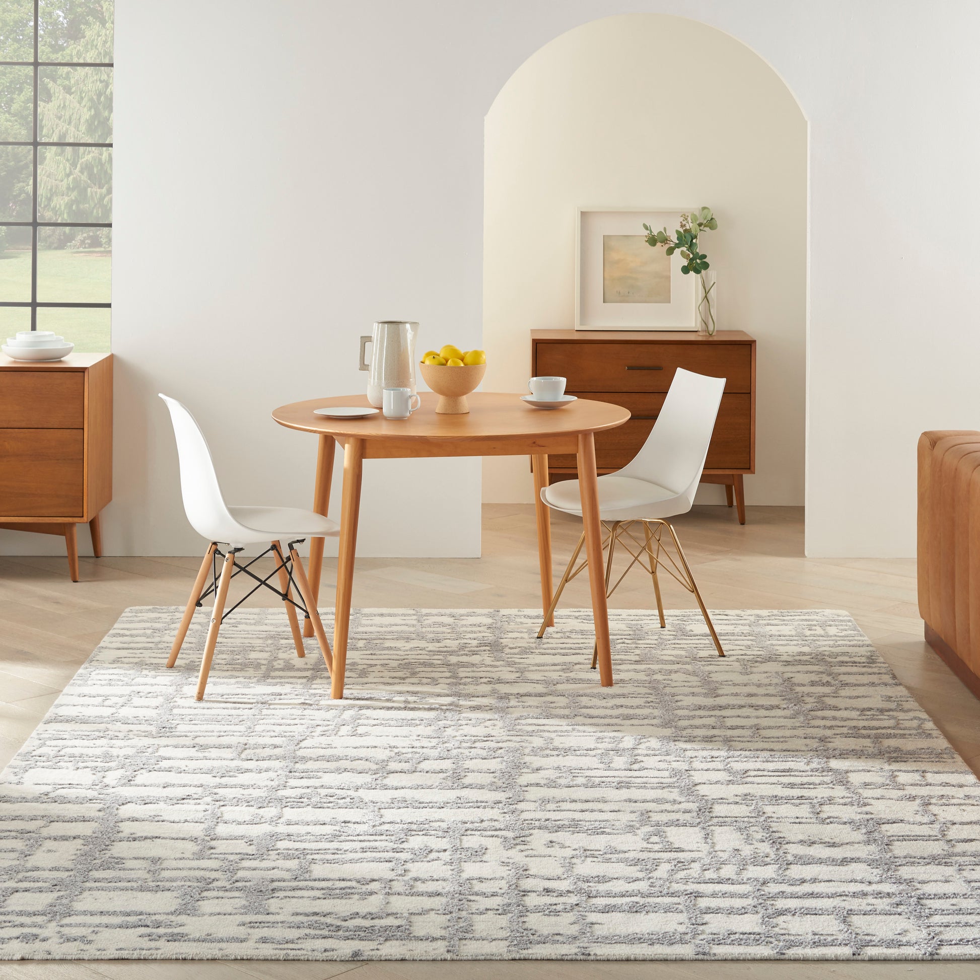 Round wooden dining table with white molded chairs on a gray patterned rug in a bright mid-century modern dining room.