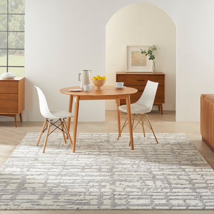 Round wooden dining table with white molded chairs on a gray patterned rug in a bright mid-century modern dining room.