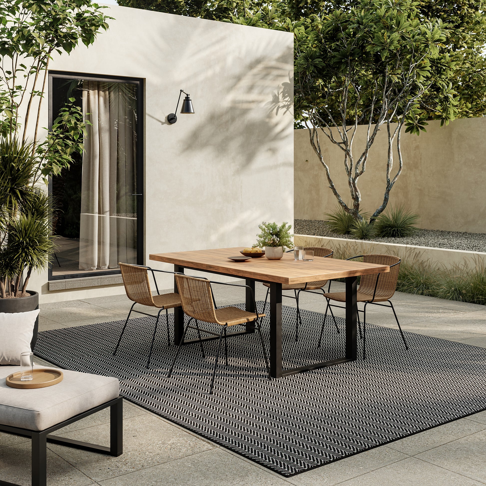 Outdoor dining area with a wood table, black metal legs, six wicker chairs on a black geometric rug.