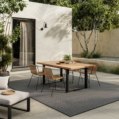 Outdoor dining area with a wood table, black metal legs, six wicker chairs on a black geometric rug.