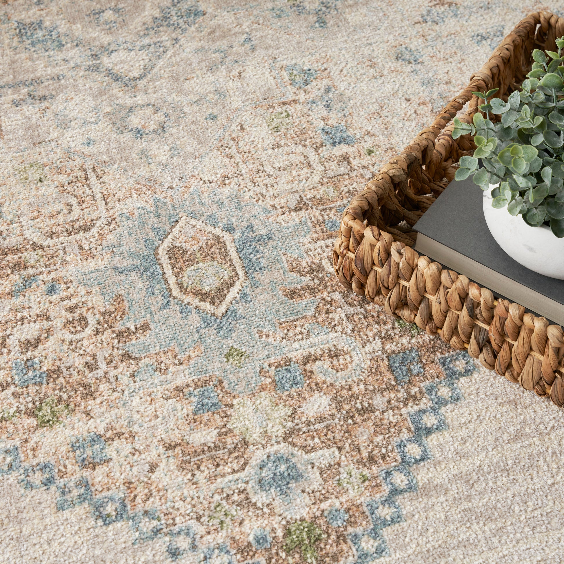 Woven wicker tray on a beige-blue geometric rug; white pot with greenery atop a gray notebook.