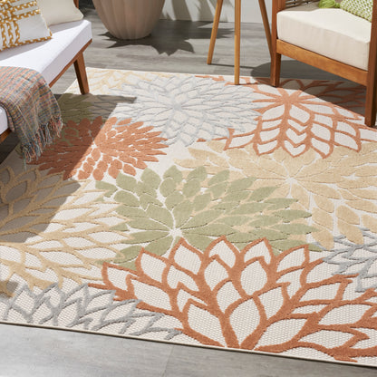 Outdoor rug with large orange, green, beige and gray leaf patterns, beside white seating and wooden chairs.