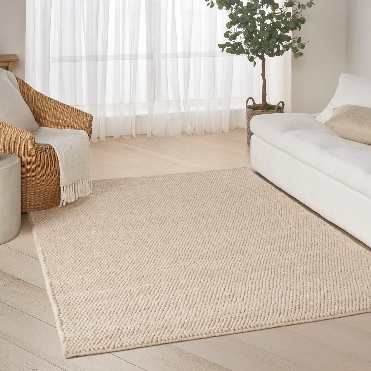Beige woven jute area rug in a bright living room with white sofa, rattan chair, plant, & sheer curtains.
