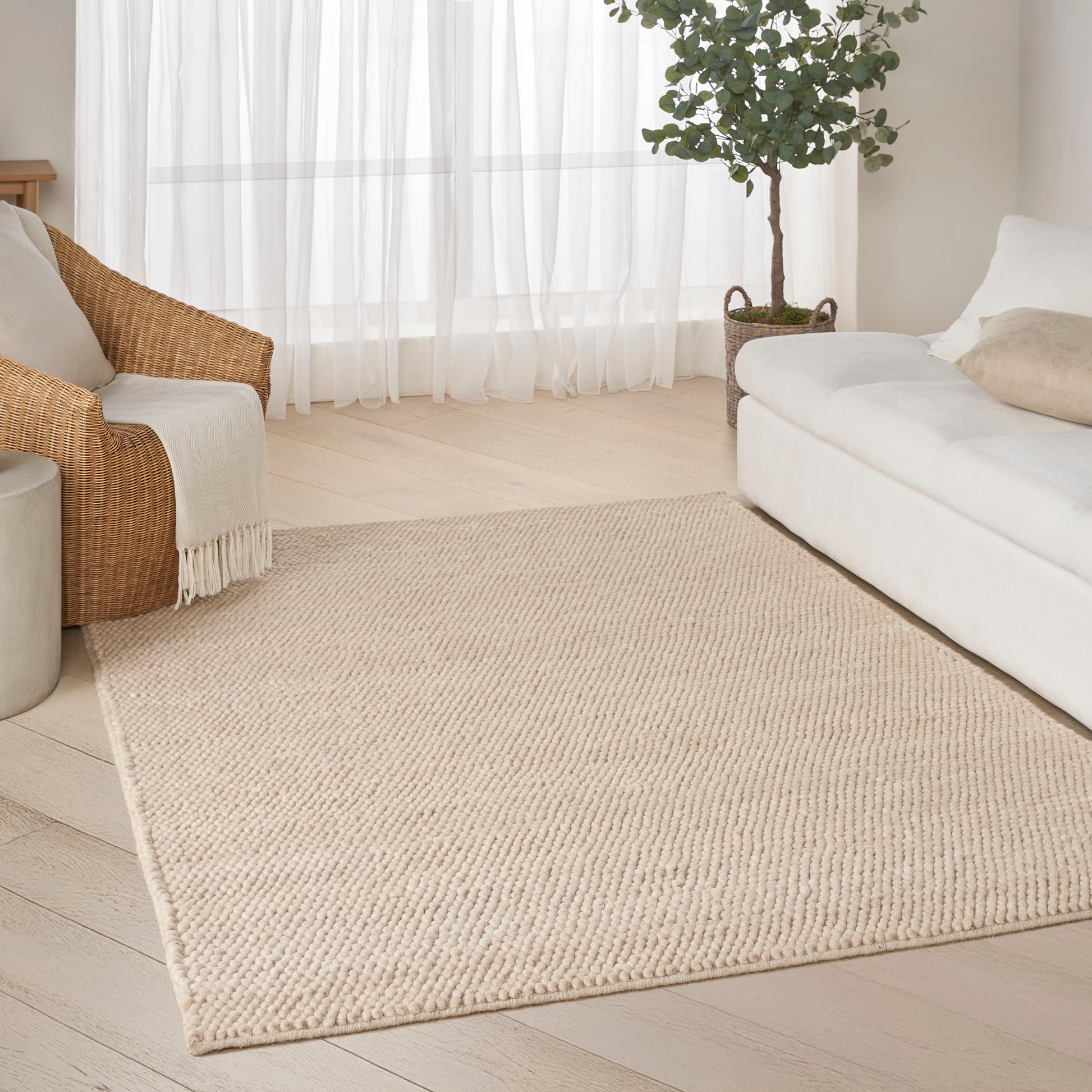 Beige woven jute area rug in a bright living room with white sofa, rattan chair, plant, & sheer curtains.