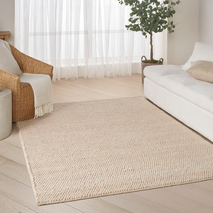 Beige woven jute area rug in a bright living room with white sofa, rattan chair, plant, & sheer curtains.